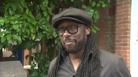 Paul Sealy, a man with long dreadlocks, wearing a large black flat cap hat, black framed glasses and a black shirt, he is standing outside his house next to the grapefruit tree 
