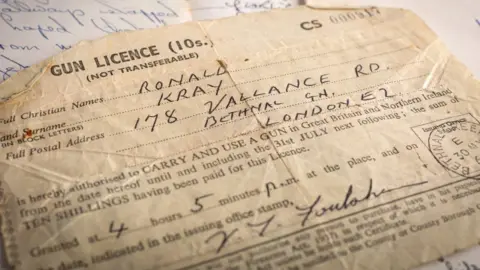 Bearnes Hampton &  Littlewood A piece of paper with the words gun licence at the top of it.  The name Ronald Kray are written in by hand and his address is given as 178 Vallance Road, Bethnal Green, London, E2.
It also has an official  date stamp of 30 May visible to the right and the signature of the official who issued the licence.
It was issued at '4 hours 5 minutes pm'.