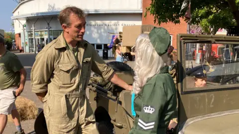 James leans with one hand on a green military vehicle, wearing a green khaki boilersuit. He is speaking to a woman with long bright blonde hair. who is wearing green 1940s costume. 