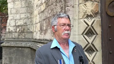 A man in a suit jacket and open-necked blue shirt stands near what appears to be a church. He has a white moustache and metal glasses. 
