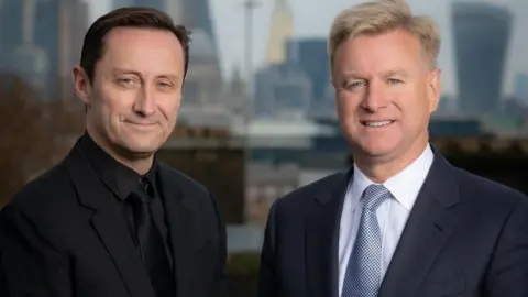 Revolution Beauty Adam is standing on the left. He has brown hair and is wearing a black suit and tie. Tom is on the right and has blonde hair and blue eyes. He is wearing a dark navy suit jacket over a white shirt and blue-spotted tie. Both are smiling to the camera.