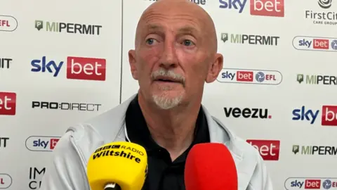 BBC Ian Holloway sitting in a press conference in front of a board with multiple advertisements of brands on. In front of him is two microphones, one is BBC Radio Wiltshire branded and the other is red. He is wearing a grey jacket and black polo, and has a goatee.