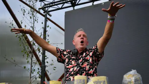 Getty Images DJ Fatboy Slim plays on stage at Glastonbury Festival. He is stood in front of a darkened screen and has his hands in the air with his mouth open. He has short grey hair and is wearing a black shirt with orange and white koi fish on it. He has a pair of headphones around his neck.