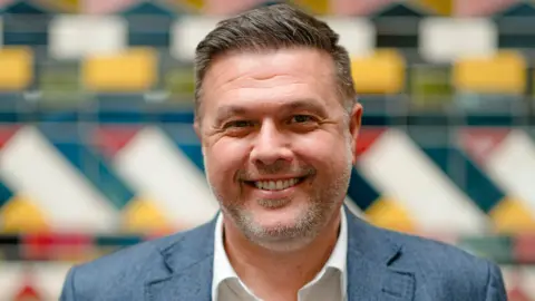 Bristol Beacon Simon smiles at the camera. He has short brown/grey hair and light grey stubble, and wears a white shirt and grey suit. He is standing in front of a blurred, multi-coloured background. 