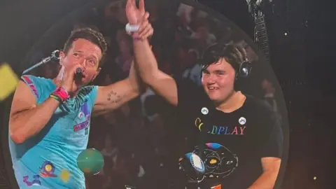 Handout Chris Martin is holding AJ Murphy's right hand in the air in this image taken from a screen at Wembley. They are sitting at a piano on stage. Martin is wearing a colourful turquoise T-shirt and colourful wrist bands. AJ is wearing a black Coldplay T-shirt and noise cancelling headphones. He is smiling.