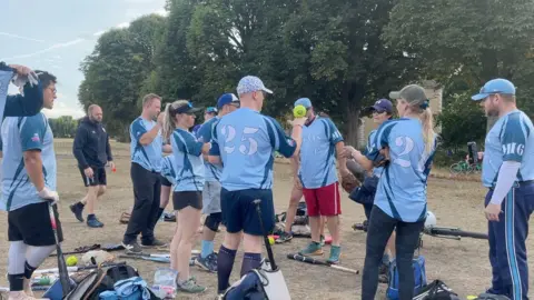BBC London Softball League team MI6 are gathered around amongst bats and other softball equipment on the grassy floor. Players, both male and female, are wearing light-blue softball t-shirts. Some are wearing dark-blue shorts, others are wearing jogging bottoms. One player, bearing the number 25 on the back of her shirt, is balancing a green softball on the back of her hand.