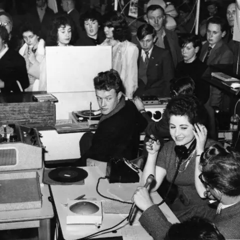 BHBN Black and white photo of the radio station with a man holding a microphone, a woman holding a phone receiver and other people in the background.