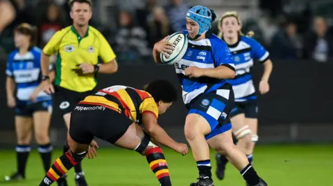 Trish Brown playing a rugby match. She is in a blue and white Bath Ladies outfit with a blue helmet. She is holding the ball and is about to be tackled by a player from the other team.