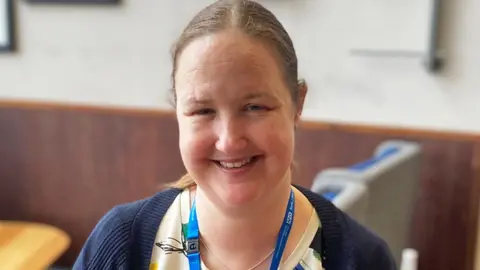 ELHT Head-and-shoulders image of a smiling Catherine Makin. She has long brown hair tied back in a ponytail with a centre parting. Smiling for the camera, she is wearing a cream top with a navy cardigan on top.
