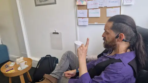 The Wallich Rob has dark hair tied back in a pony tail and a beard. He is wearing a purple shirt which is open at the collar, a black waistcoat and a large silver pendant necklace. He is sitting in his office and chatting. He is to the right of the photo, above him is a notice board with various white notes on it. There is a coffee table in front with a white mug, which has a cup of tea in it. 