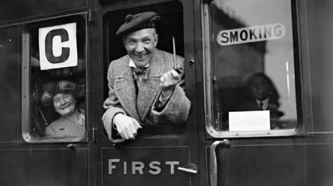 PA Media Sir Harry Lauder, a very jolly elderly man with a pipe and lowland bunnet, is hanging out the window of a 1920s railway carriage. A woman in a broad hat and tweed coat is smiling at his antics from inside the train carriage.