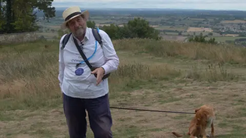 The Cotswold Explorer Robin Shuckburgh talking to the camera, while holding his dog on a leash. It is a cloudy day. He is wearing a white T-shirt in support of Sepsis Research.