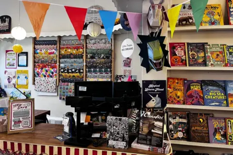 The cashier's desk at a book store. There is a dark brown desk, with a red and white striped decoration on the side. A computer screen is on right of the desk, surrounded by books including 'Resistance' and 'Magical Unicorn Friend'. There is a small gap on the desk to the left of the computer. To the left of this is a poster which reads 'Help us save Moon Lane Books', with a QR code. There is a display of wrapping paper on the white wall behind the desk, and multi-coloured triangle banners are displayed from the ceiling. To the right of the desk, there is a set of shelves with colourful children's books on display.