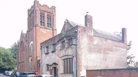 A large re brick church building, with cars parked in front and greenery behind it