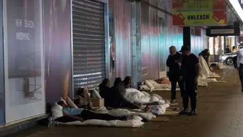 Facundo Arrizabalaga/MyLondon Several people are sleeping rough on the pavement under a building's awning. An advert saying "UNLEASH" hangs above them and the shop's shutters are down. 