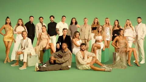 Rachel Joseph/Objective Entertainment The cast of The Only Way is Essex in front of a green backdrop. Twenty-one of the cast are standing and the rest are sitting either on green seats or on the floor.