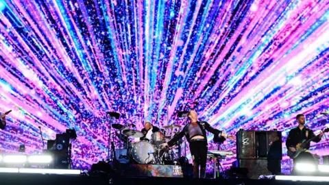 Coldplay frontman Chris Martin is stood on the front of a stage leaning backwards and dancing, wearing a black jacket and black trousers with a purple-red tye-dye T-shirt. He is in front of a large screen, which has bright pink and blue lines radiating from the middle. Bassist Guy Berryman is playing on the right in a black jacket, with drummer Will Champion playing at the back of the stage behind Chris Martin.
