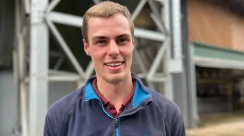 A man with short brown hair smiling at the camera. He is wearing a jumper with a maroon shirt underneath.
