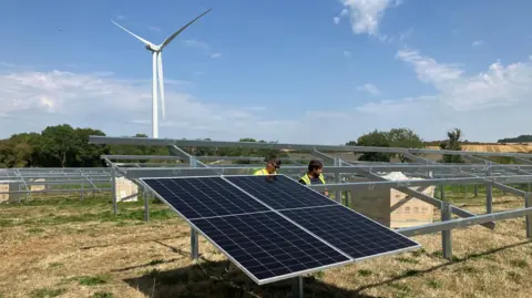 Amy Holmes/BBC A picture of two men installing solar panels. They are both wearing yellow high visibility jackets and there is a wind turbine behind them and it is a sunny day with blue skies. 