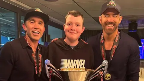Sarah Waller Aiden Waller stands in front of a large national league trophy in between Ryan Reynolds (right) and Rob McElhenney (left). All three of them smile at the camera. Both Ryan and Rob wear Wrexham black caps and dark long sleeve t-shirts, while Aiden, who has short dark hair, wears a black Marvel hoodie. 