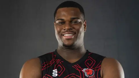 Essex Police/Essex Rebels Basketball Anthony 'TJ' Hibbert in a black basketball vest with a red pattern and Essex Rebels logo. He smiles at the camera in front of a grey background.