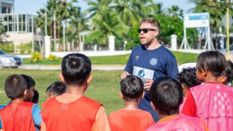 Marshall Islands Soccer Federation A group of young boys wearing orange and pick sports vests over their t shirts are getting instruction from a their coach. He is wearing sunglasses and there are palm trees in the background