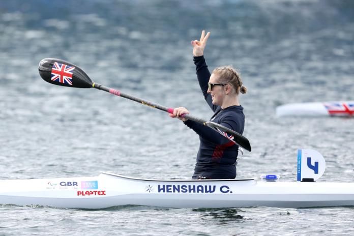 A woman in a canoe holds her oats with one hand and lifts the other hand in the air, making a peace sign