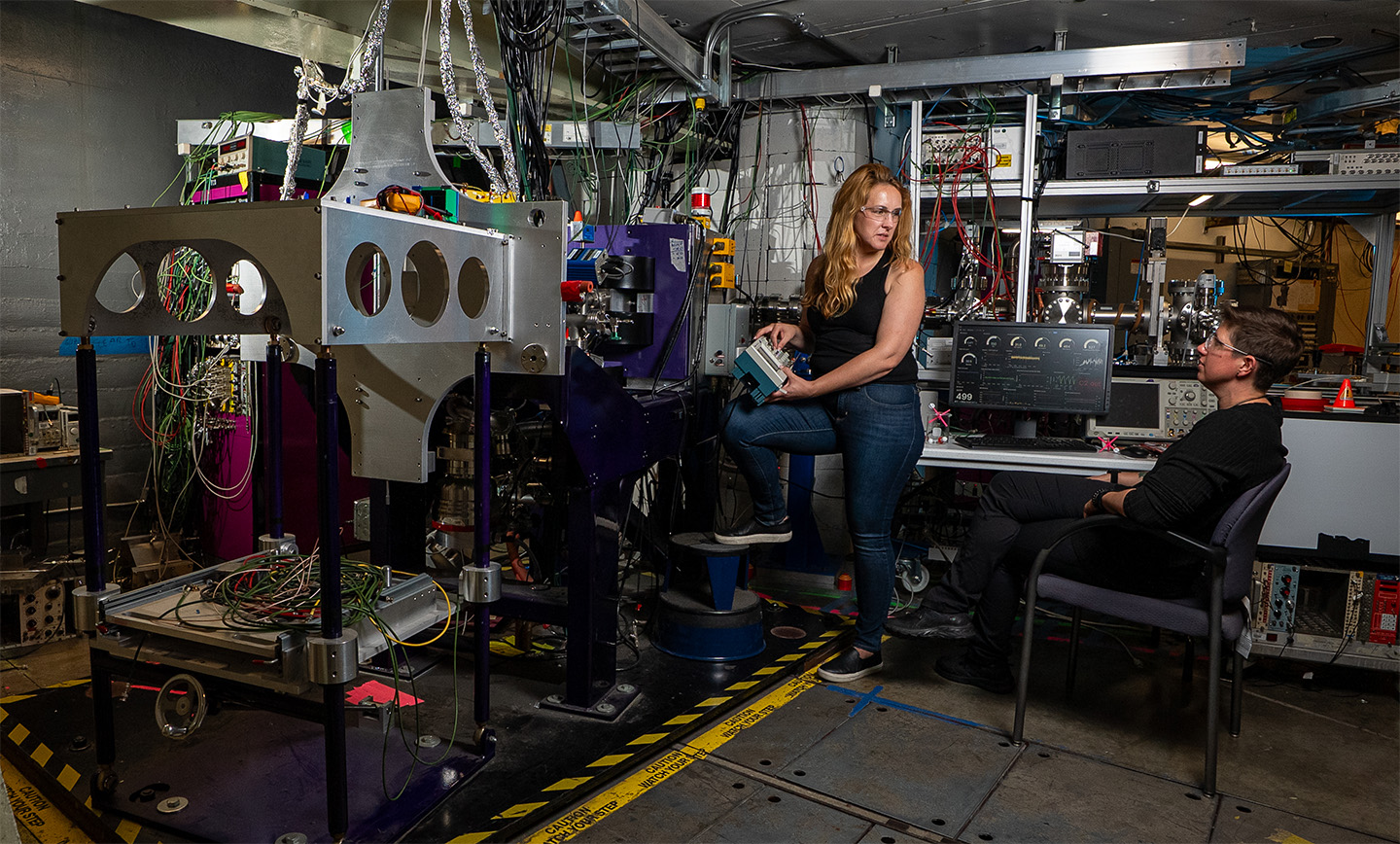 Jennifer Pore stands holding a piece of equipment and is talking to Jacklyn Gates, seated at a computer. The lab equipment of the FIONA detector setup is prominent.