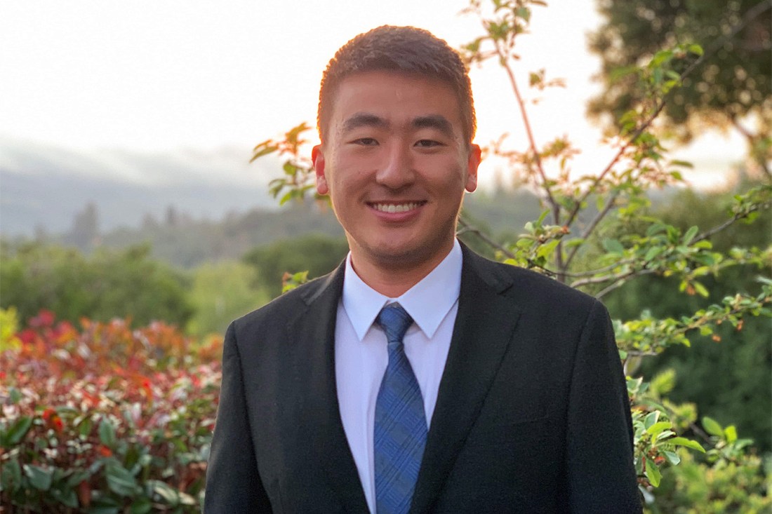 Portrait of Norman Yuan standing outside in front of greenery wearing a suit and tie.