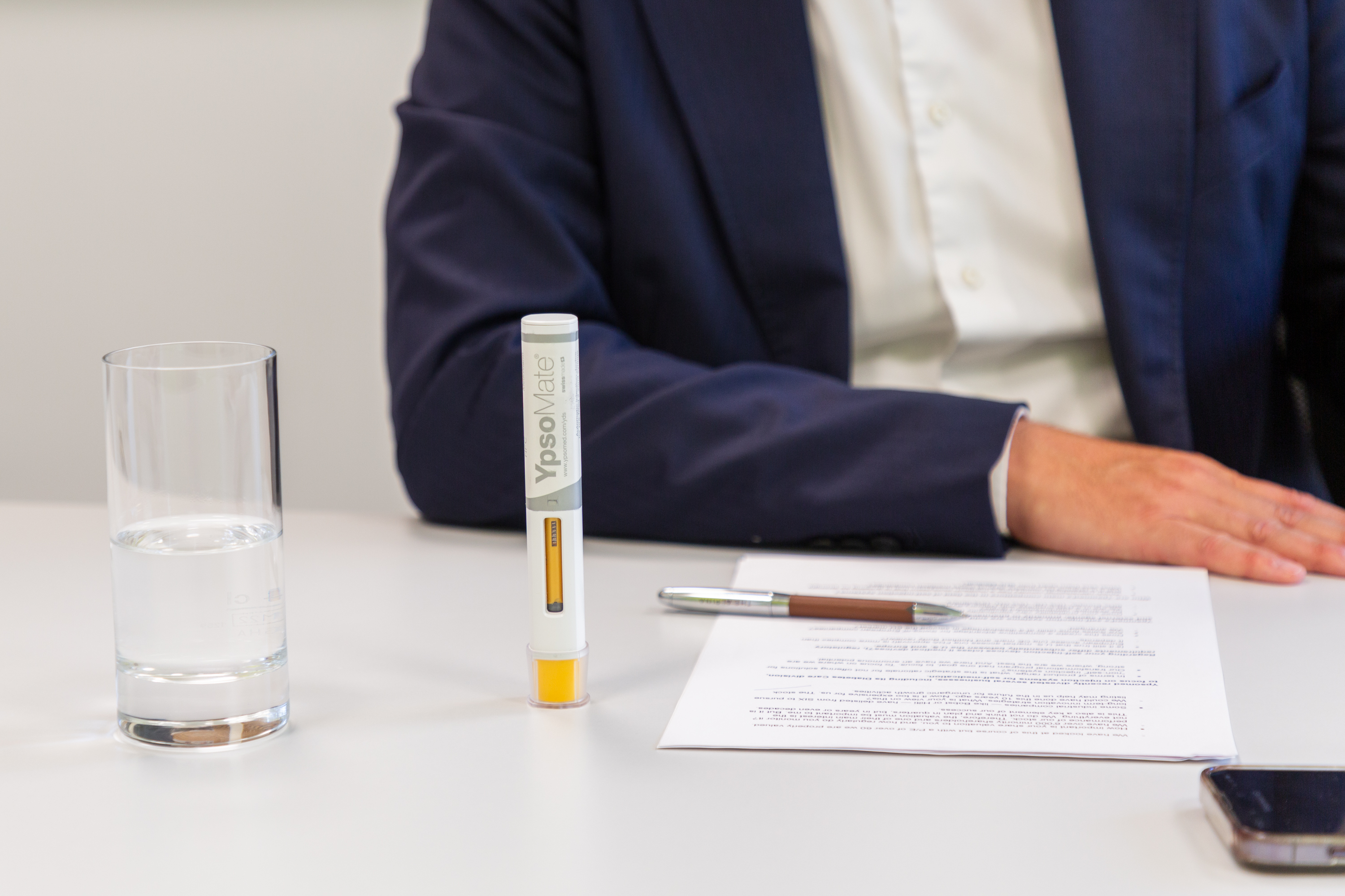 A person sits at a table with documents, a pen for self-injection, a pen and a glass of water in front of them.