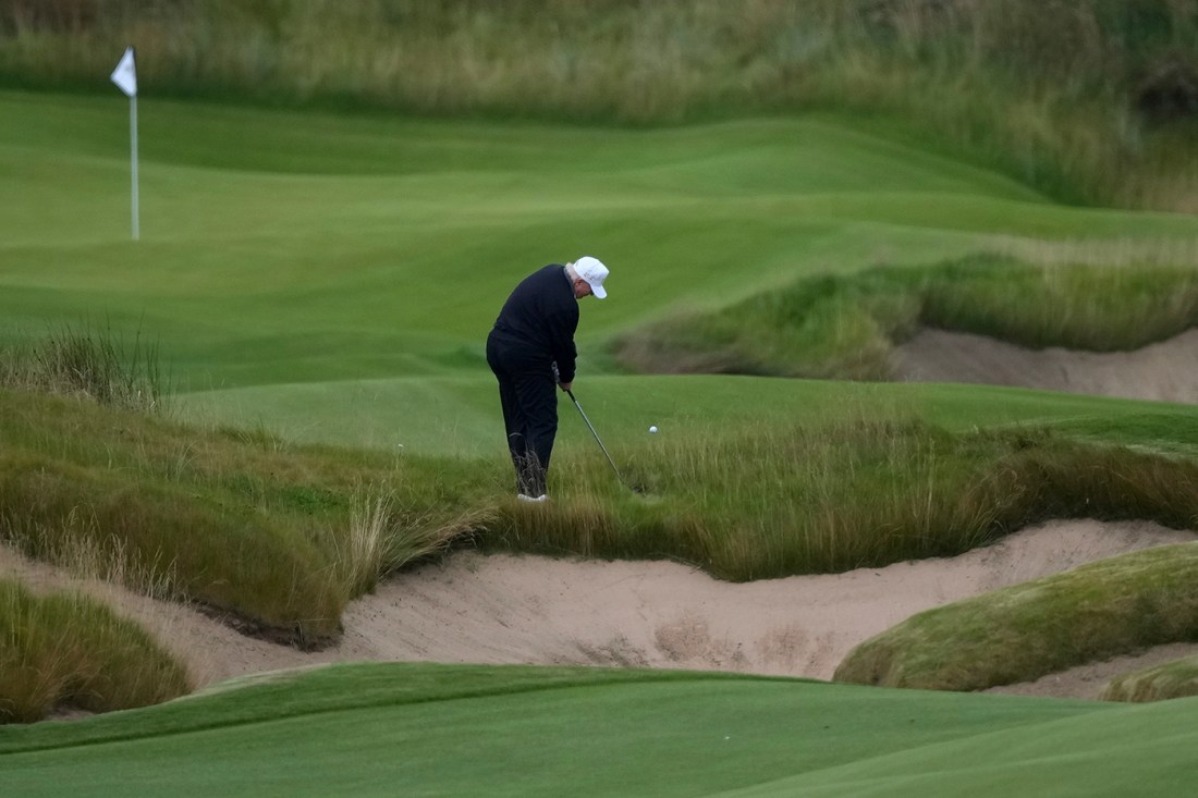 President Donald Trump wearing all black clothing and a white baseball cap playing golf.