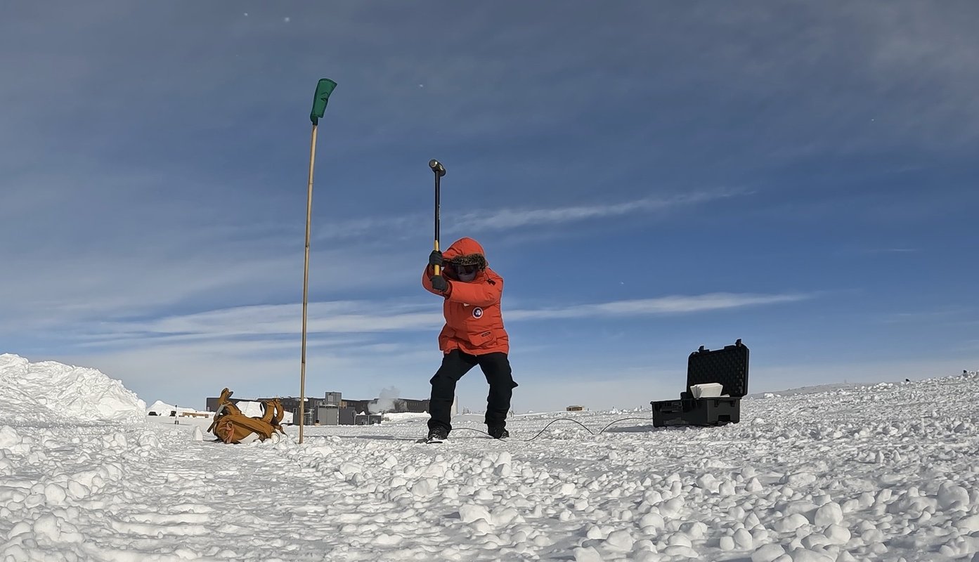 Zhongwen Zhan in a bright red winter coat swinging a sledgehammer at the Antarctic ice