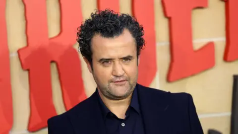 EPA Daniel Mays is at the premier of The Thursday Murder Club in London. The title of the film is written behind him. He has olive skin and is wearing a dark blue coloured blazer and shirt.