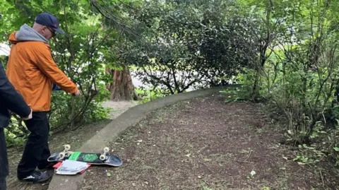 Dr Kenny Brophy A man in an orange jacket and baseball cap pointing at an area of grass and dirt, where a skatepark used to be