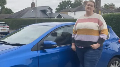 BBC Mary Bates is standing in front of her blue car outside her home.