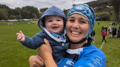 Trish Brown Trish Brown in a blue rugby shirt and blue helmet smiling, holding her baby son, who is also smiling and wearing a blue hoody. They are on a rugby pitch.