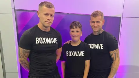 A 10-year-old boy standing in between two men who have muscular and tattooed arms. The three are all wearing black T-shirts that read "Boxwave" and they are all smiling at the camera.