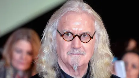Getty Images Billy Connolly, with long grey hair, a grey goatee and small round glasses, looks into the camera
