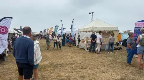BBC Gazebos in rows. There are people walking around the gazebos. There are flags next to the gazebos. The grass is green and brown. The sky is grey and full of clouds. 