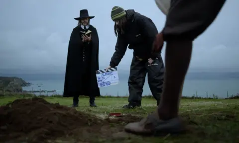 Andrew Butler An actor standing in a field on a cliff dressed in a historical black puritan's outfit and hat, holding up a small book. A member of the film crew holds up a clapper board saying: Albert. Another actor's shoe and leg are in view.
