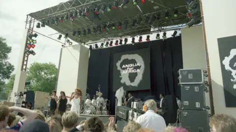 Mark Hodgson A wide shot of an outdoor concert stage seen in daylight, with a large logo at the back of the stage reading 'Anglia for Africa', over a shape of Africa inside an outline shape of Norfolk and Suffolk. Performing on the stage are three women in glamorous dresses. The backs of the heads of some of the crowd can be seen at the bottom of the frame, as can a camera operator looking into his camera as he focuses it on the group.