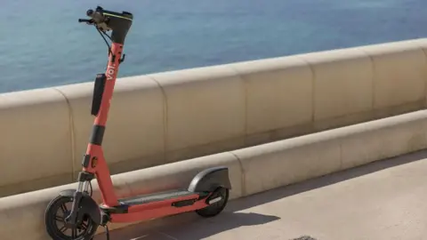 Voi An orange e-scooter parked on a pavement and resting against a concrete sea wall. Beyond the wall is blue sea.