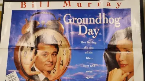 Hansons A film poster for Groundhog Day. It features the name of the main actor Bill Murray along with the title of the film. Underneath, it reads: "He's having the day of life life...over and over again." On the left is a cock with Billy Murray's face in the middle. On the left is the actress Andie MacDowell.

