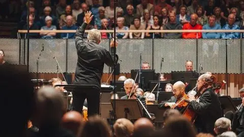 Giulia Spadafora A conductor, dressed in black, raises one hand in the air mid-performance in front of a strings sections. From a raised balcony, an audiences watches the musicians play. 