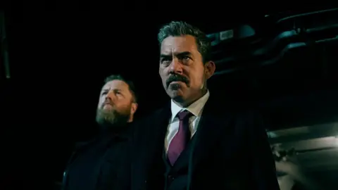 Pat Redmond Two men are looking forward. It's dark.
The man in the foreground has greying short hair and a moustache.
He is wearing a dark coat, over a dark suit. His shirt is white and his tie purple.
The man behind is dressed fully in black. He has lighter hair and a full beard