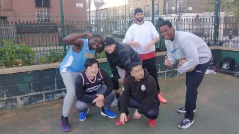 Ray Akpofure A group of six men on a basketball court. They are wearing sports clothes, and are clustered together, with some of them crouching down.