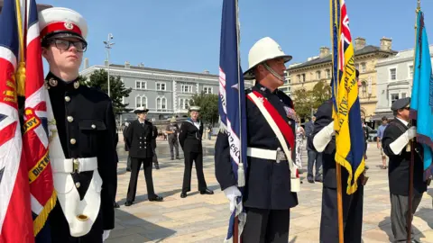 BBC Four men in military uniform stand in a line in a town square, each holding a different flag. Members of the Navy can be seen standing behind them. There are grand buildings at the edge of the square.
