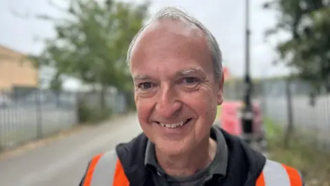 Jasmine Lowe/BBC A man with short grey hair is smiling into the camera for a head and shoulders shot. He is wearing orange high vis, a black jumper and polo shirt. Behind him is an alleyway with blurred metal fences on both sides.
