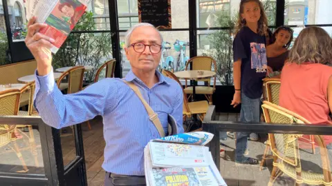BBC Ali Akbar holds aloft a copy of Le Monde in front of a café in central Paris