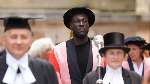 PA Media Stormzy is in the middle of a crowd, which is blurred, walking towards the camera. He is wearing a black T-shirt and suit jacket, and has a flat doctorate hat on and a pink and red gown. 